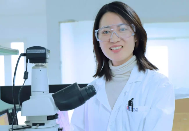 A smiling scientist in protective eyewear stands beside a microscope in a lab, representing CGT ophthalmology research and development.