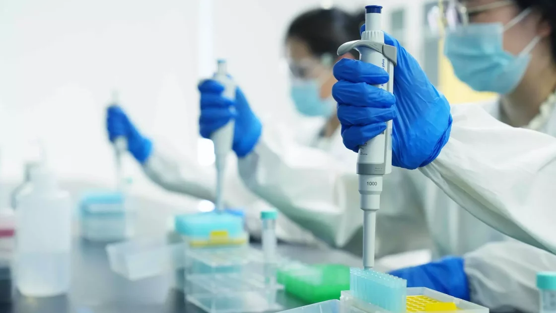 Researchers in a lab wearing protective gear and blue gloves focus on pipetting samples for large molecule biologics analysis, surrounded by lab equipment and reagent bottles.