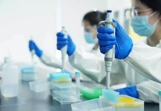 Researchers in a lab wearing protective gear and blue gloves focus on pipetting samples for large molecule biologics analysis, surrounded by lab equipment and reagent bottles.