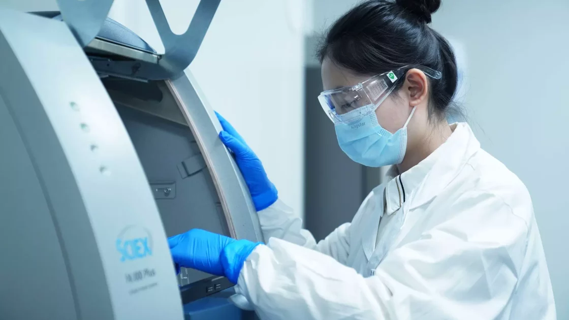 A scientist wearing a lab coat, protective eyewear, a face mask, and blue gloves is operating a SCIEX 7500 mass spectrometer in a laboratory environment. The individual is focused on the equipment, performing a radiolabelled ADME​ scientific procedure for bioanalytical development.