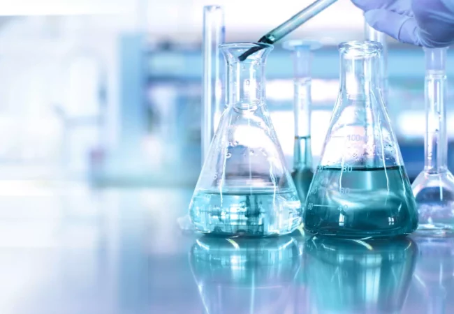 A scientist uses a pipette to add liquid into a glass flask, with other flasks filled with blue solution on a reflective lab surface.