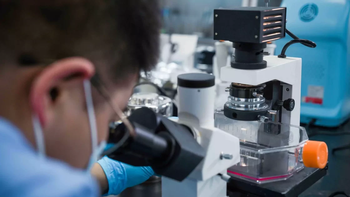 A scientist wearing a mask and gloves uses a microscope in a lab, performing a CGT safety assessment with specialized equipment.