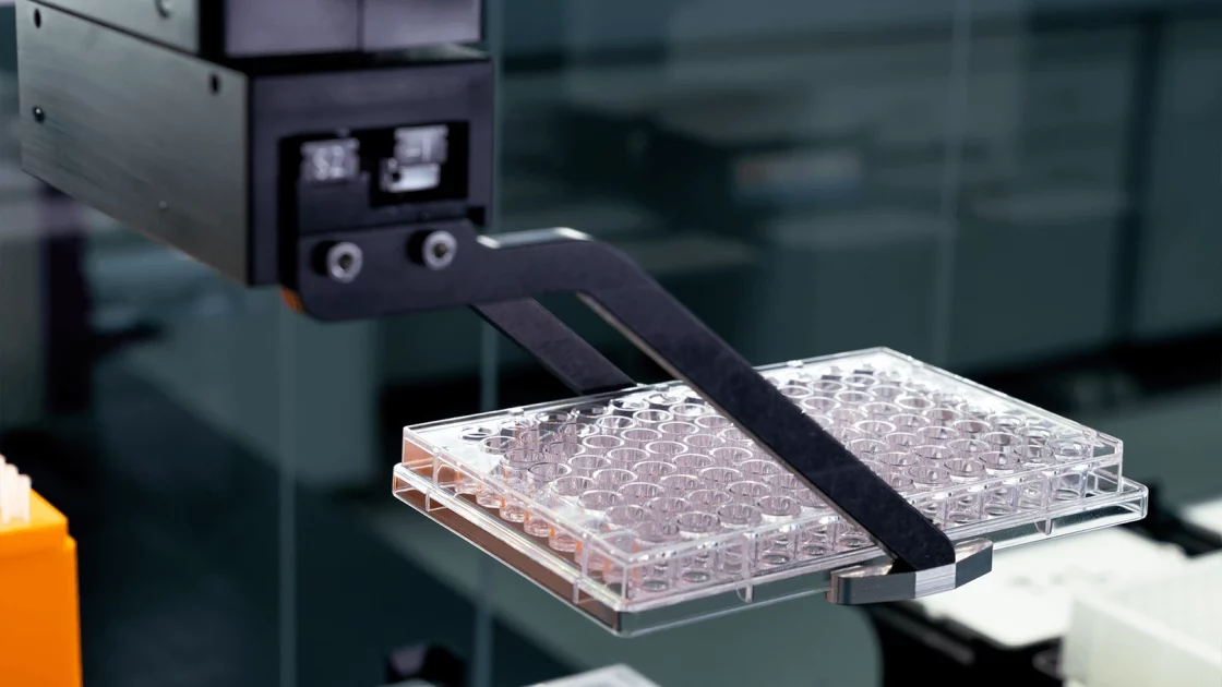 A robotic arm holds a 96-well plate used for drug metabolism and pharmacokinetics (DMPK) studies in a laboratory setting focused on automated analysis.