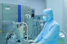 A scientist in full-body protective gear works at a biologics CDMO (Contract Development and Manufacturing Organization) lab, operating an ÄKTAprocess system from Cytiva. The sterile environment and advanced equipment highlight large-scale biologics production, such as purification and filtration of therapeutic proteins.