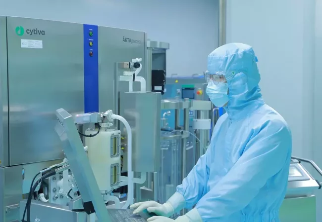 A scientist in full-body protective gear works at a biologics CDMO (Contract Development and Manufacturing Organization) lab, operating an ÄKTAprocess system from Cytiva. The sterile environment and advanced equipment highlight large-scale biologics production, such as purification and filtration of therapeutic proteins.
