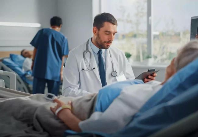 A doctor reviews a patient’s chart while another patient rests in the background in a hospital room, with a focus on human AME studies.