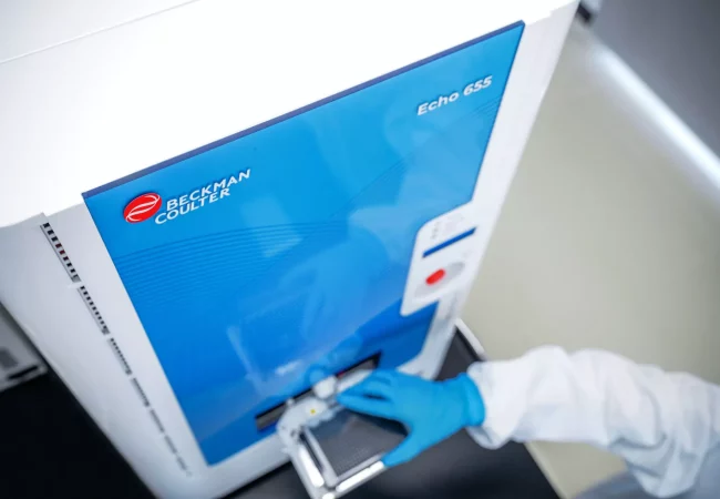 Scientist operating a Beckman Coulter Echo 655 device, used for automation in in vitro biology workflows, with gloved hand loading a plate.