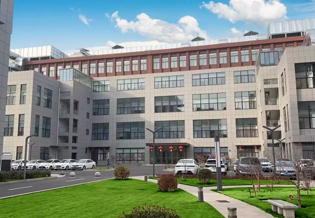 Qingdao office building with large windows, surrounded by green lawns and pathways. Several parked cars are visible in front of the building, which houses cutting-edge labs focused on preclinical ADME studies and bioanalytical development. The sky is clear with a few clouds.