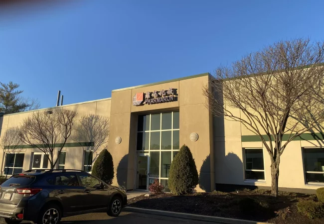 A single-story building with large windows and the Pharmaron logo above the entrance, located at the Woburn site, with trees and a parked car in front.