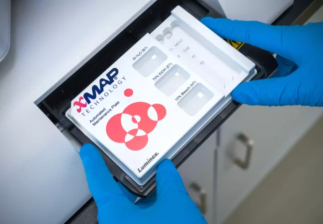 A scientist inserts an xMAP Technology automated maintenance plate into a Luminex system, wearing blue gloves. The plate contains solutions like DI water, ethanol, and bleach, used for system calibration and upkeep.