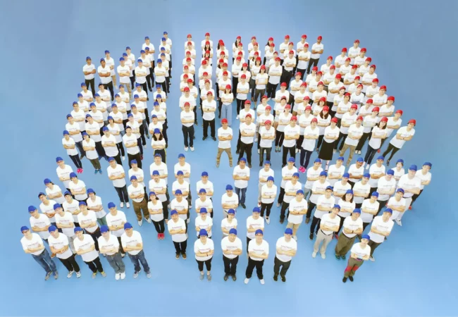 Pharmaron management team posed against a blue background, wearing coordinated blue and red hats. Their arrangement forms the shape of the Pharmaron logo icon, showcasing team unity and branding.
