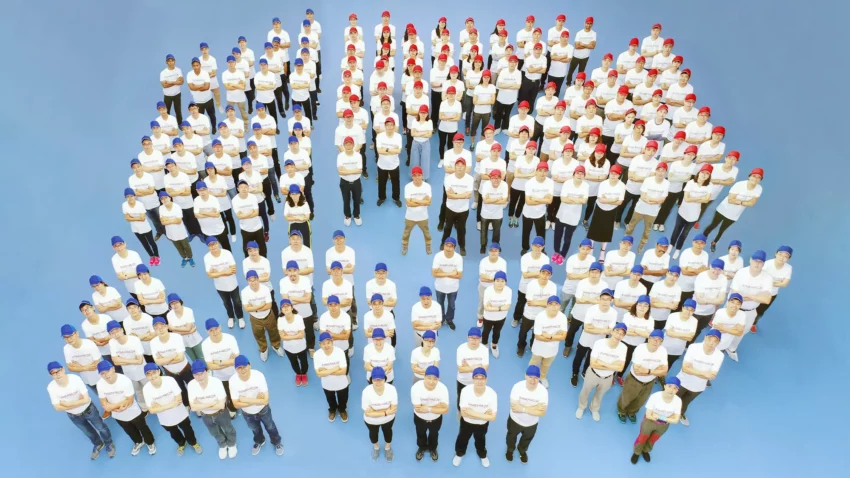 Pharmaron management team posed against a blue background, wearing coordinated blue and red hats. Their arrangement forms the shape of the Pharmaron logo icon, showcasing team unity and branding.