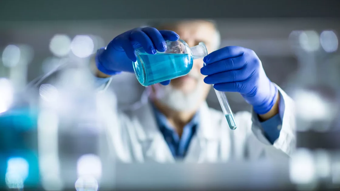 A scientist wearing blue gloves carefully pours blue liquid from a flask into a test tube in a laboratory setting. The background is blurred, emphasizing the precise handling of chemicals. in radiosynthesis and radiolabelling services for ADME