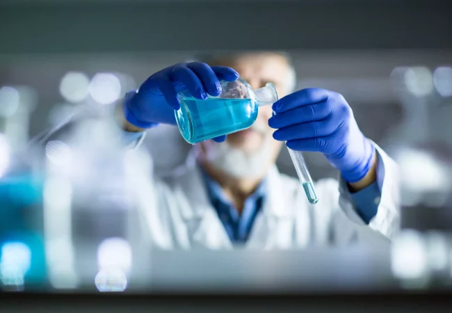 A scientist wearing blue gloves carefully pours blue liquid from a flask into a test tube in a laboratory setting. The background is blurred, emphasizing the precise handling of chemicals. in radiosynthesis and radiolabelling services for ADME