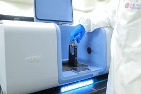 A scientist operates a Bruker instrument, placing a sample into the device while wearing protective gloves and lab attire, preparing for analysis.