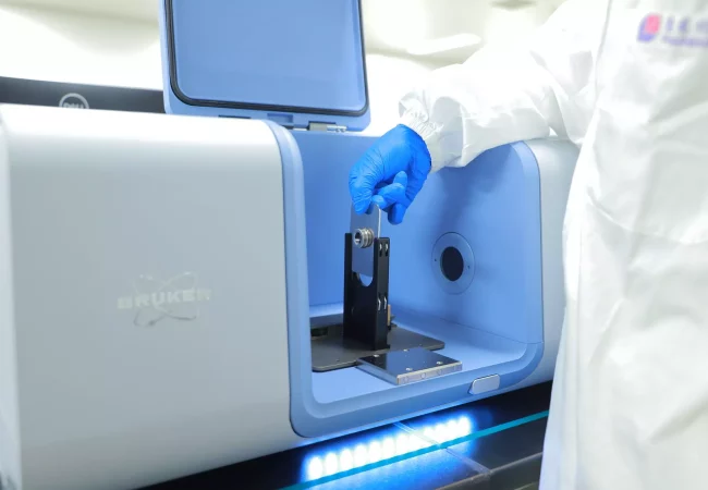 A scientist operates a Bruker instrument, placing a sample into the device while wearing protective gloves and lab attire, preparing for analysis.