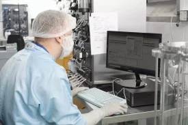 A scientist in a sterile GMP manufacturing facility works at a computer, monitoring data from bioprocessing equipment. The complex system of tubing and valves suggests a CDMO setup for large-scale biologics production, ensuring compliance with Good Manufacturing Practices (GMP).