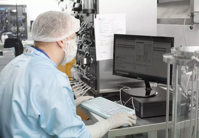 A scientist in a sterile GMP manufacturing facility works at a computer, monitoring data from bioprocessing equipment. The complex system of tubing and valves suggests a CDMO setup for large-scale biologics production, ensuring compliance with Good Manufacturing Practices (GMP).