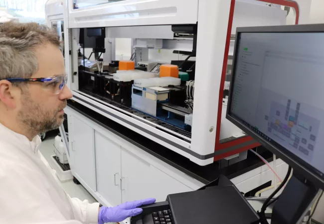 A scientist monitors a high-throughput process development (HTPD) robot through a computer interface. The automated system is equipped with various modules for handling samples, optimizing biopharmaceutical production processes in a laboratory setting.