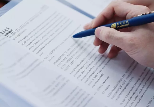 A hand holding a Pharmaron-branded pen reviews detailed documents, reflecting quality assurance protocols and quality control standards in the pharmaceutical industry, forming the foundation for a quality management system