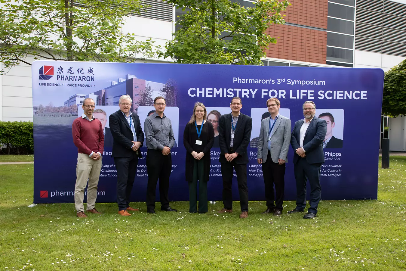 Group of scientists posing in front of a banner for Pharmaron's 3rd Symposium, "Chemistry for Life Science," with a backdrop of a research facility.