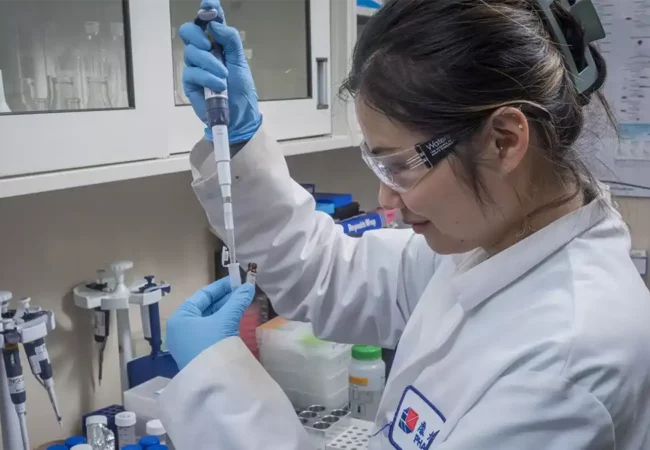 Scientist in a Pharmaron lab coat using a pipette to transfer liquid into a vial, wearing gloves and safety glasses in a well-equipped laboratory.