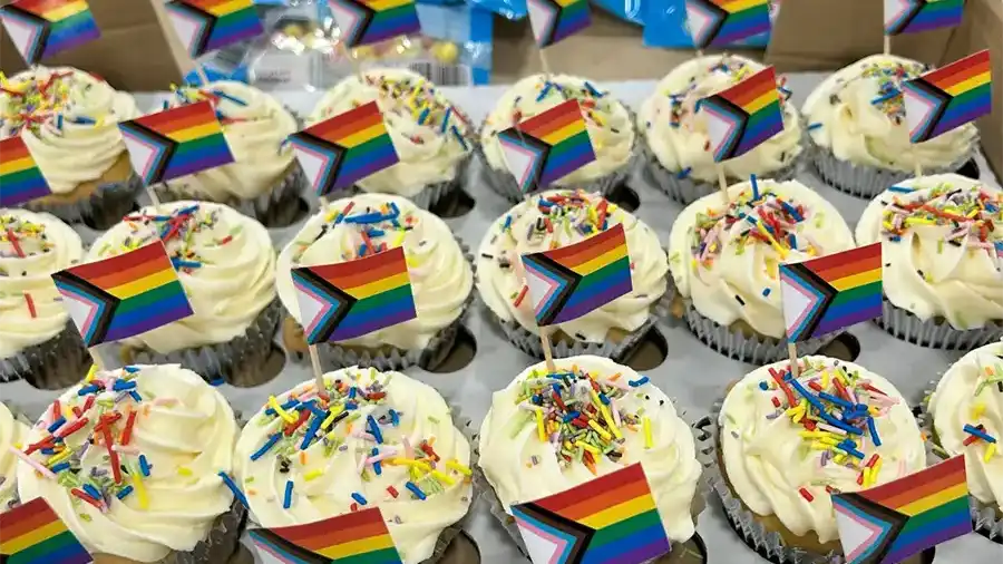 Cupcakes topped with rainbow sprinkles and small Progress Pride flags arranged in rows for a Pride-themed celebration.