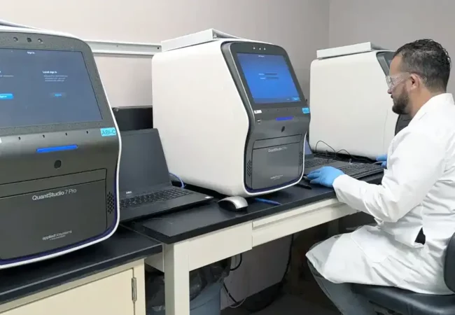 A scientist in a white lab coat and gloves working with a QuantStudio 7 Pro instrument in a molecular biology lab.