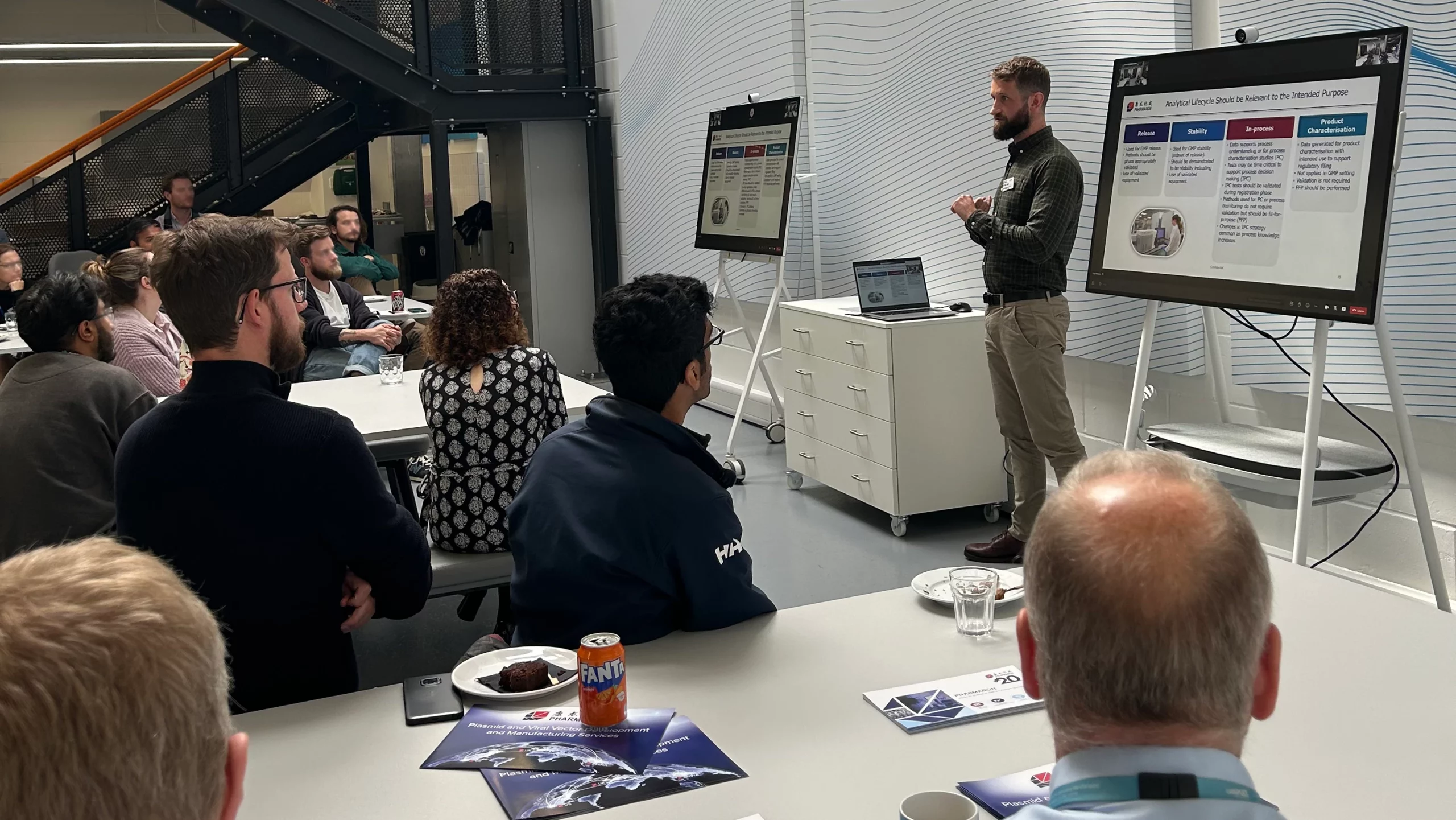 Scientist presenting CMC strategies on analytical lifecycle to a seated audience during a Lunch & Learn event, with slides displayed on screens.