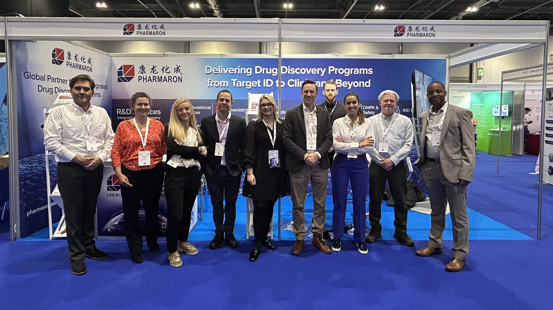 Pharmaron team members stand together in front of a booth promoting drug discovery programs at a professional conference, smiling and wearing event badges.