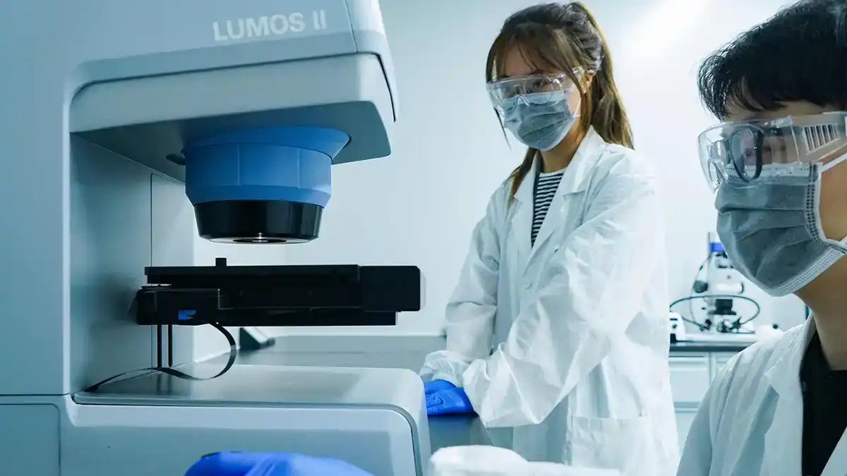 Two scientists in protective gear use a Lumos II instrument for analytical testing, supporting comprehensive developability assessment services.