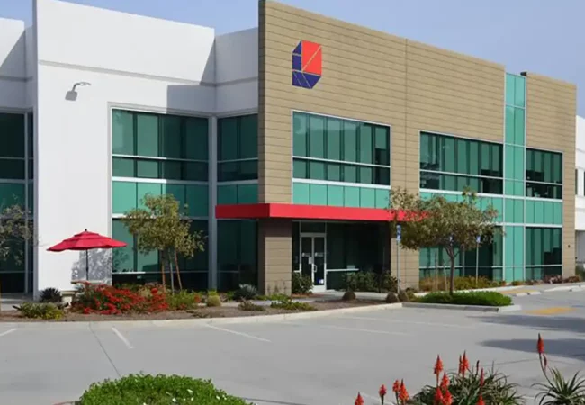 Modern two-story Pharmaron facility in San Diego with large glass windows, a red entrance awning, and an outdoor seating area with red umbrellas.