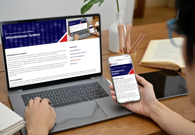 A person viewing Pharmaron's Whistleblower System webpage on both a laptop and smartphone, with open books and stationery nearby on a wooden desk.