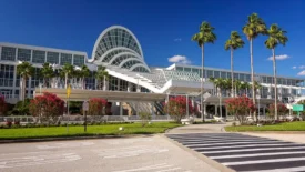 Wide-angle view of the Orange County Convention Center under a clear blue sky, framed by tall palm trees and vibrant red flowering shrubs. A crosswalk leads towards the building's modern glass architecture and arched skylights.