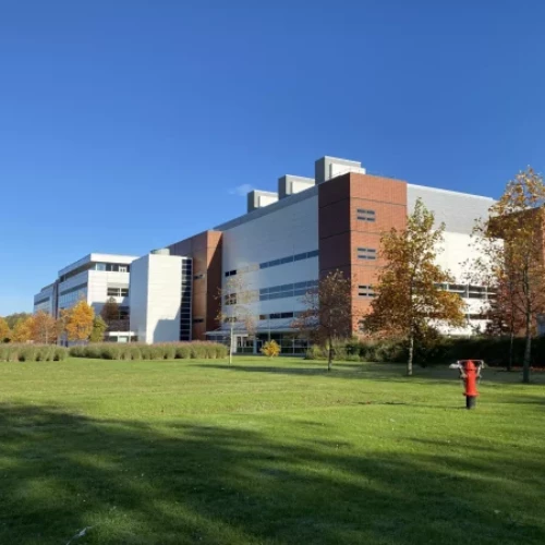 Pharmaron's Hoddesdon site, a modern multi-story facility with red and white exterior, surrounded by green lawns and autumn trees under a clear blue sky.