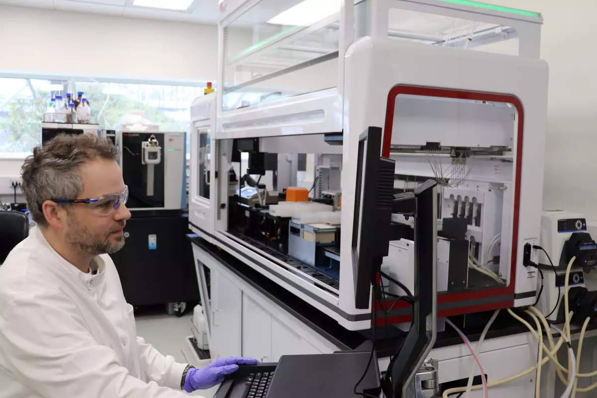 A scientist wearing safety goggles and gloves works at a computer next to advanced laboratory equipment in a well-lit laboratory environment. The equipment includes automated machinery with visible components and tubing, reflecting a high-tech research setting.