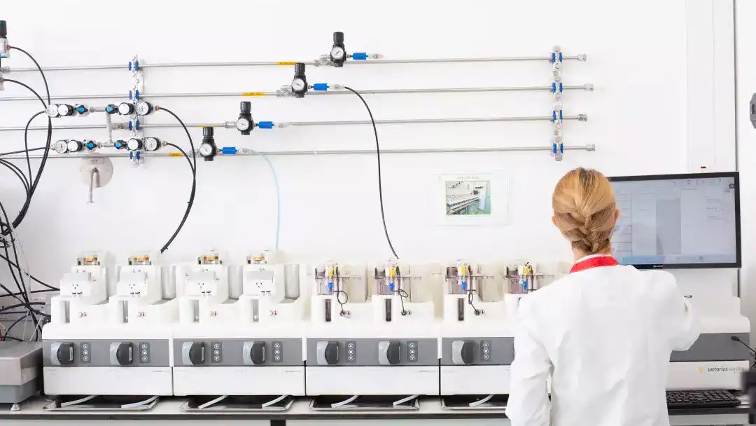 A scientist wearing a lab coat operates a AMBR250 bioreactor system in a laboratory setting. The instrument is connected to a computer displaying analytical data, with lab equipment.
