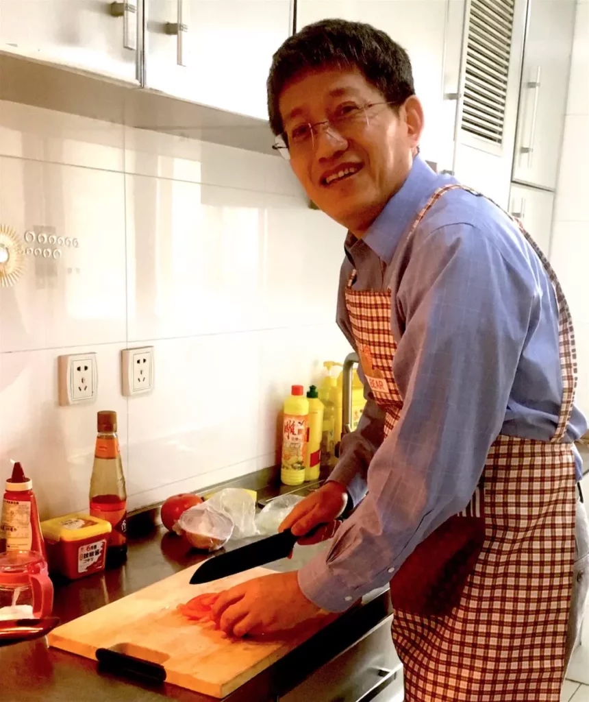 A smiling man in glasses and a checkered apron slices tomatoes on a wooden cutting board in a well-lit kitchen with various condiments on the counter.