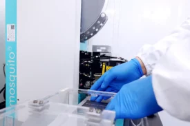 Scientist wearing blue gloves operates a Mosquito crystallization robot in a cleanroom setting for structural biology lab services.
