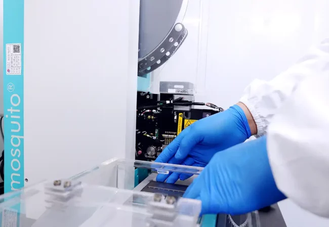 Scientist wearing blue gloves operates a Mosquito crystallization robot in a cleanroom setting for structural biology lab services.