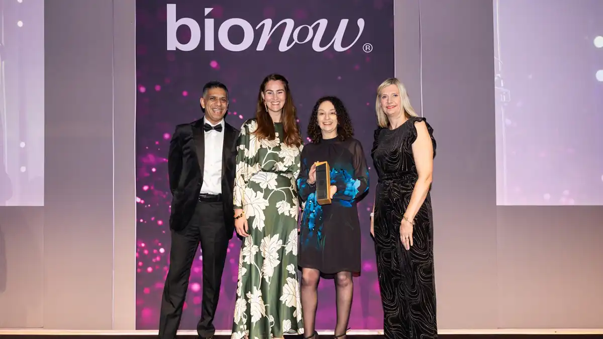 Four people stand on stage at the Bionow Awards, smiling as one holds a golden trophy, with a sparkling purple backdrop displaying the "Bionow" logo.