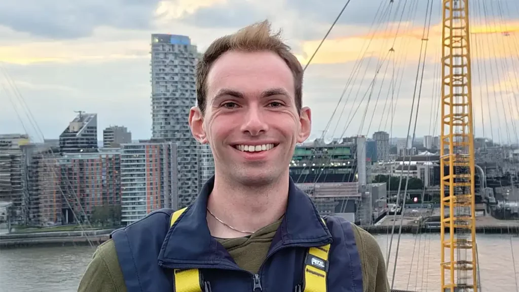 Smiling man in safety harness stands on rooftop at sunset with modern London skyline and O2 Arena cables in background.