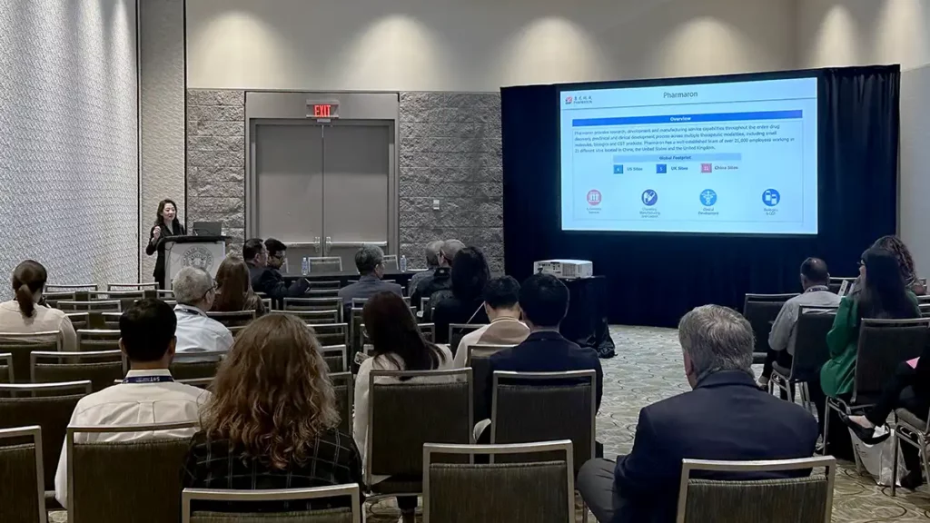 Scientist presents Pharmaron overview slide to a seated audience in a conference room, highlighting global sites and service capabilities.