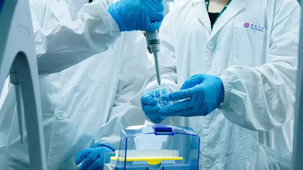 Scientists in Pharmaron lab coats use a pipette to transfer liquid into a beaker, wearing blue gloves in a sterile environment.