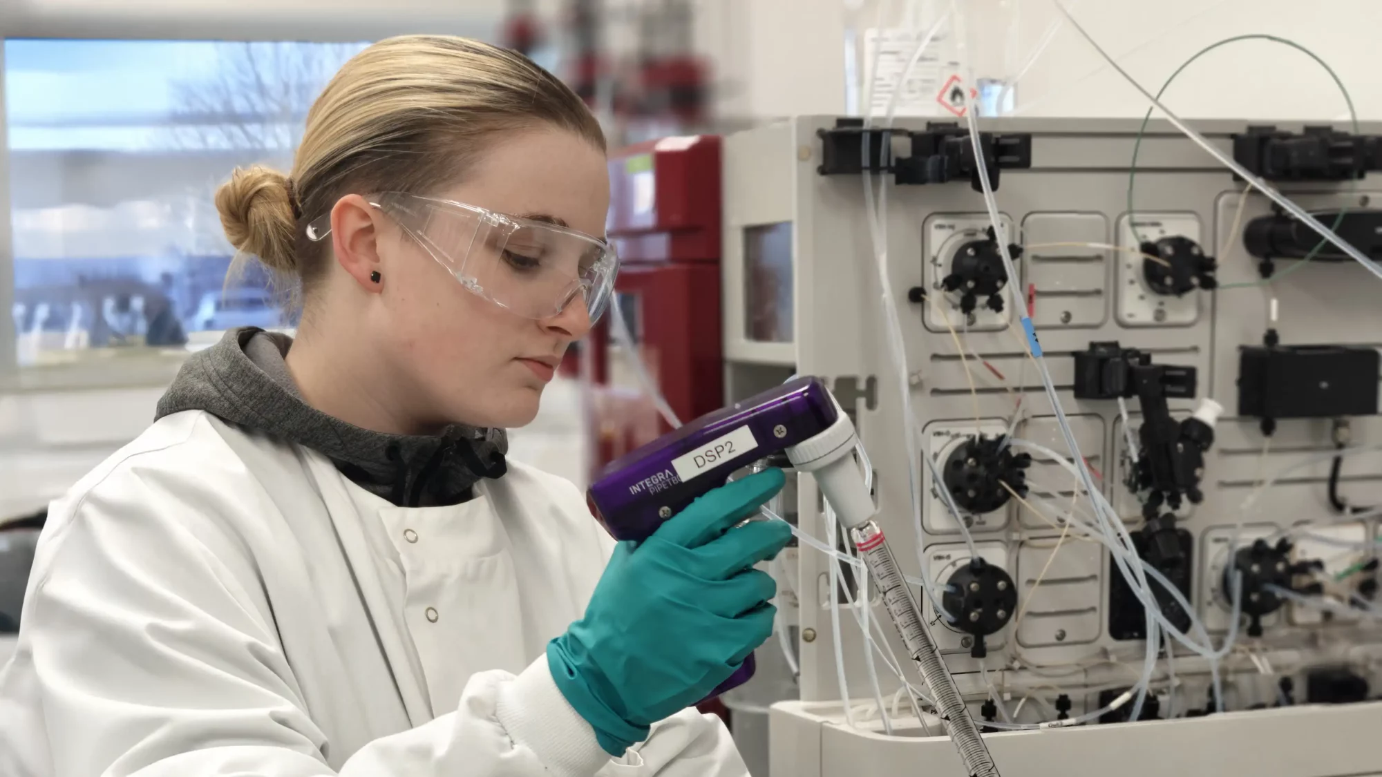 Scientist in lab coat and gloves uses a pipette controller labeled DSP2 near a complex bioprocessing instrument.