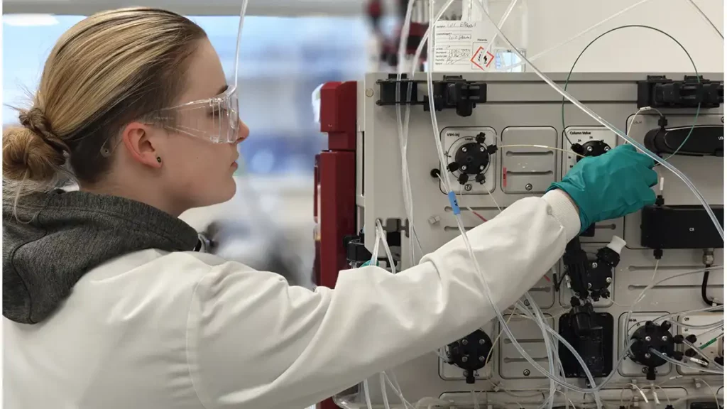 Scientist wearing safety gear adjusts tubing on a bioprocessing system with multiple valves, gauges, and transparent lines.